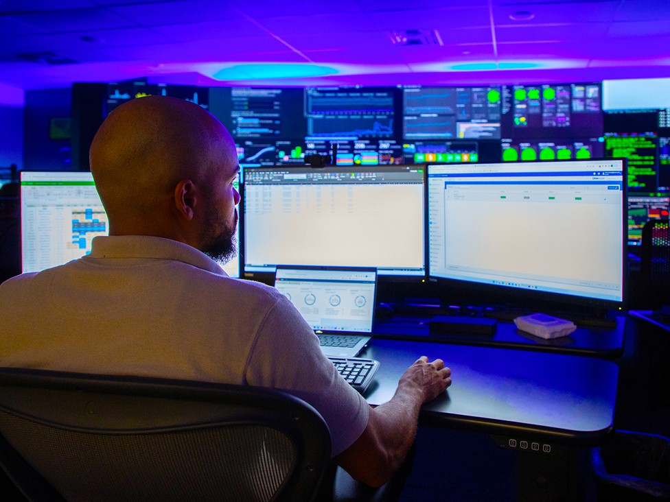 J.B. Hunt employee sitting at a desk with three monitors and a laptop displaying charts and statistics in a room with walls covered in screens with blue lighting