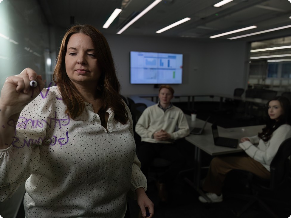 employees in a conference room discussing strategy while one writes on a glass wall