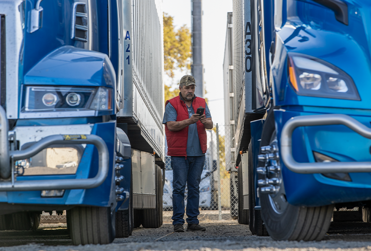 A truck driver stands between two semi trucks and looks at their phone